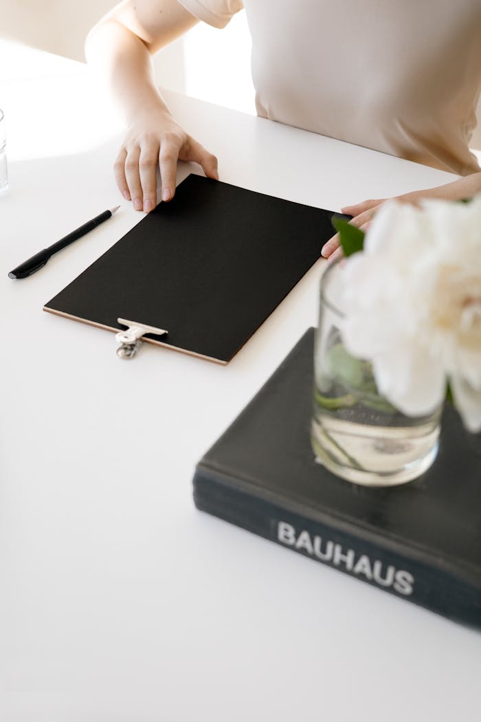 Aesthetic minimalist workspace featuring a clipboard, book, and flowers on a clean white desk.